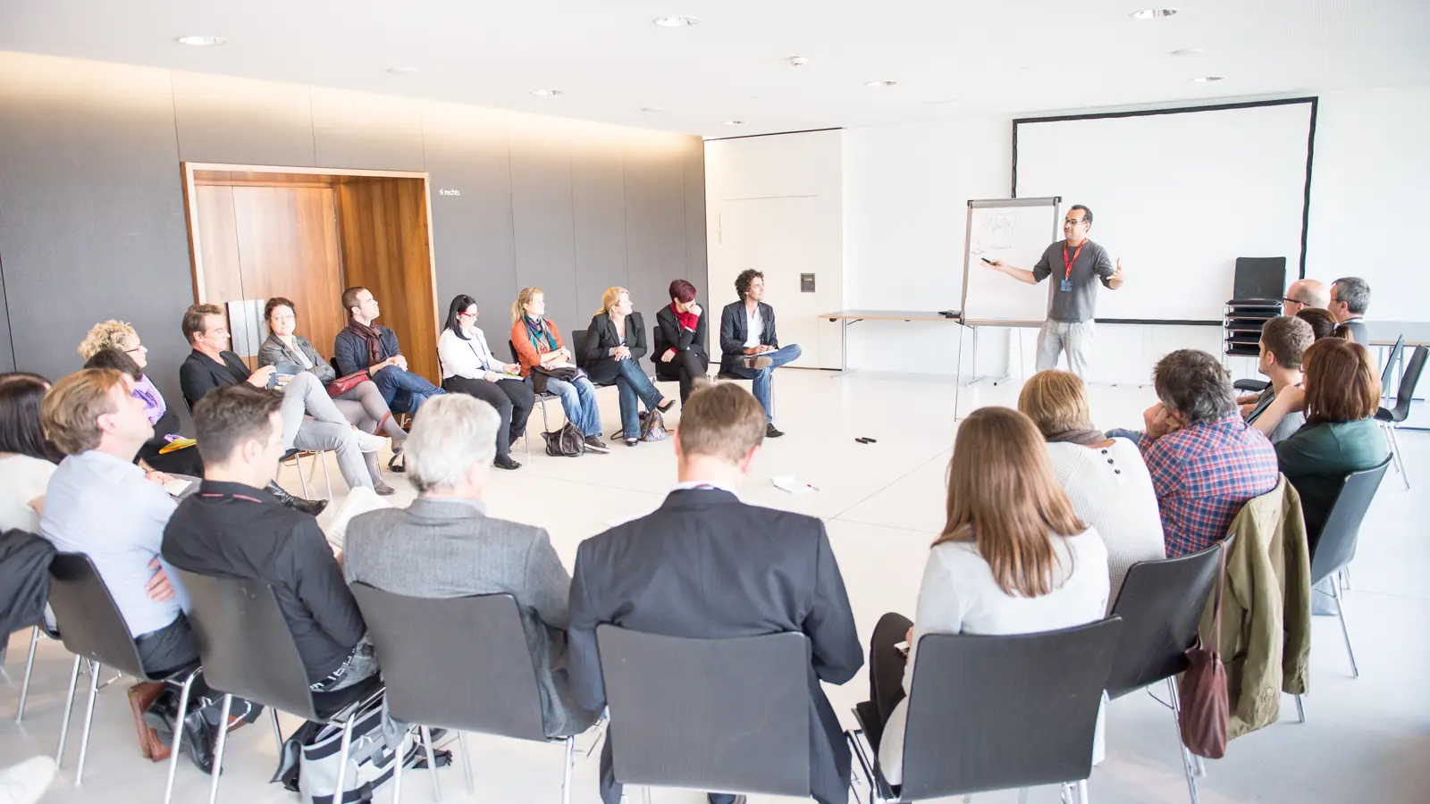 Workshopteilnehmer:innen in einem Stuhlkreis und einem Vortragenden mit Flipchart und Leinwand in der weißen Seegalerie © Matthias Rhomberg