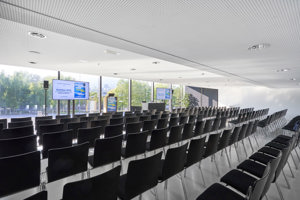 Schwarze Stuhlreihen vor zwei Großbildschirmen im weißen Saal Bodensee im Festspielhaus Bregenz mit Blick auf den Vorplatz