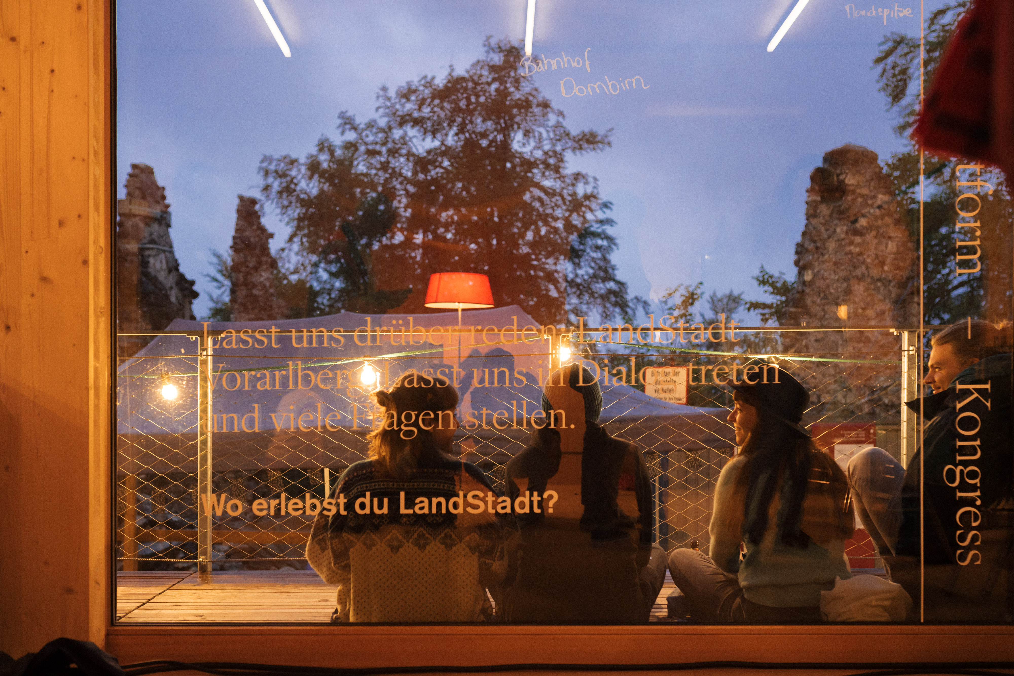 LandStadt Vorarlberg. Drei junge Menschen sitzen auf einer verglasen Terrasse © Hanno Mackovitz